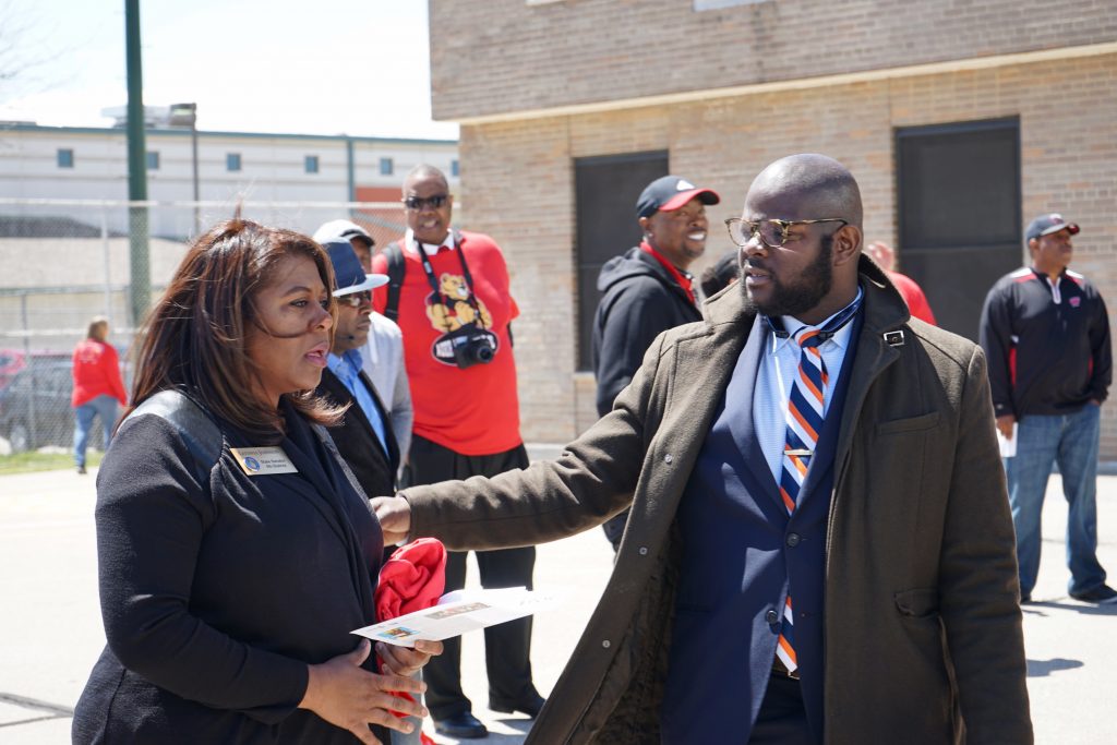 People stand outdoors near a brick building. One person in a coat and striped tie gestures toward another person holding papers, while others gather in the background.