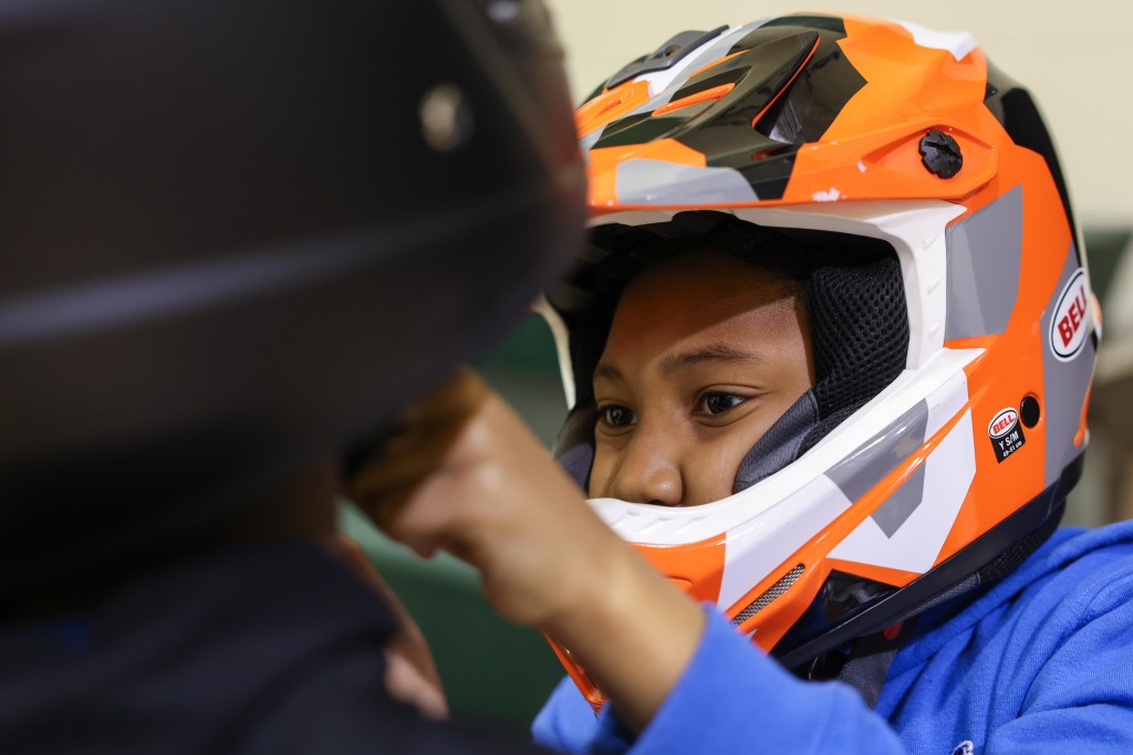 A person wearing an orange and white full-face helmet and a blue top looks to the left and adjusts the helmet of another person.