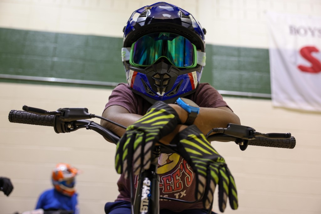 A person wearing a full-face helmet and goggles rests with crossed, gloved hands on bike handlebars inside a gym, with another helmeted rider blurred in the background.