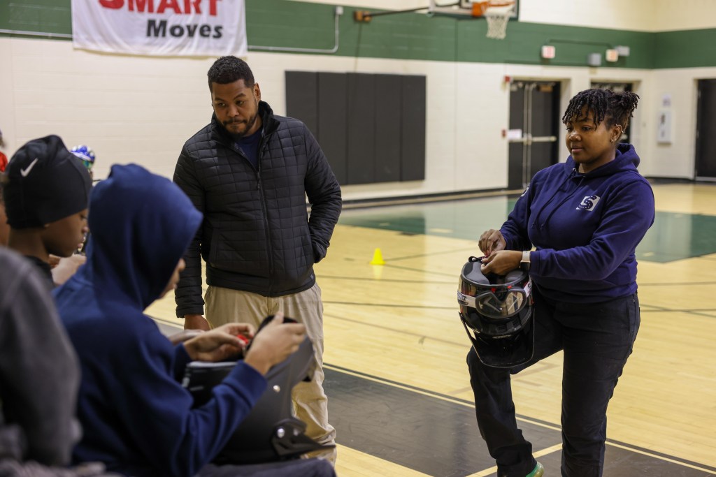 Two people stand on a gym floor as one of them holds a helmet, looking at other people who are sitting, with a banner reading “SMART Moves” on a wall in the background.