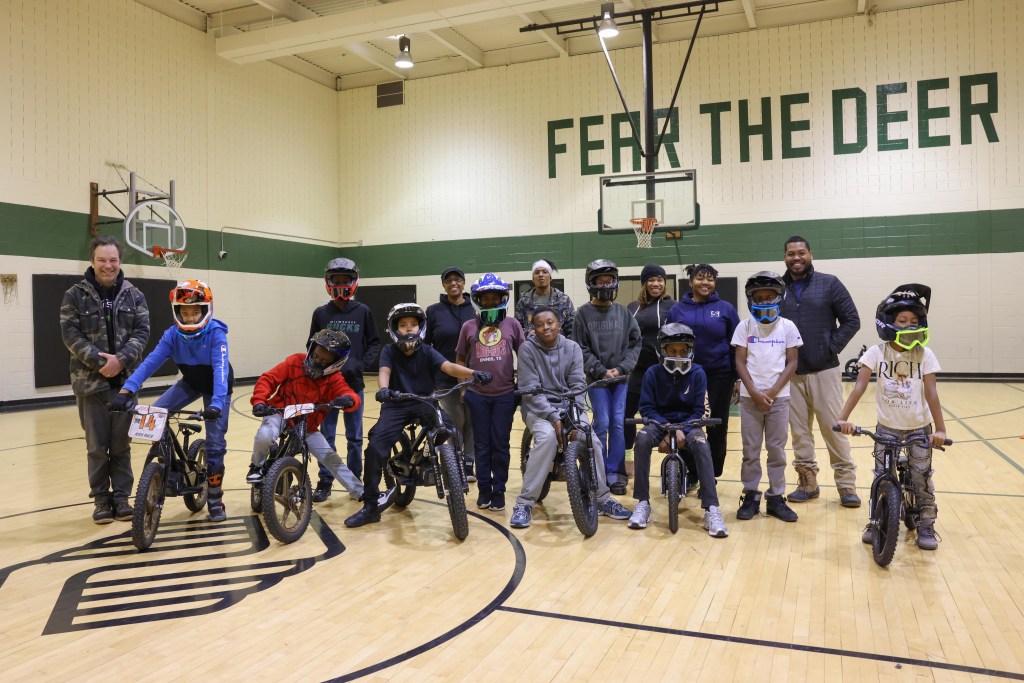 People, some of them wearing helmets, stand and sit with bicycles on a gym floor beneath two basketball hoops, with “FEAR THE DEER” on the wall behind them in green letters.