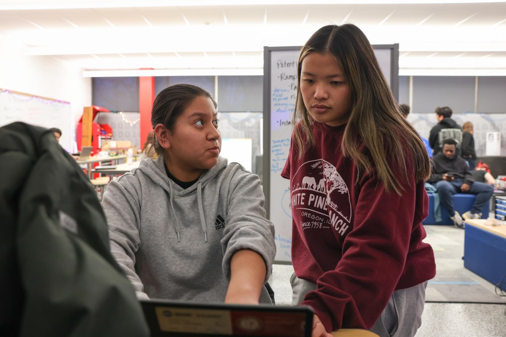 Two people are next to a laptop, with one pointing at the screen, inside a room with blue seating and a whiteboard behind them.