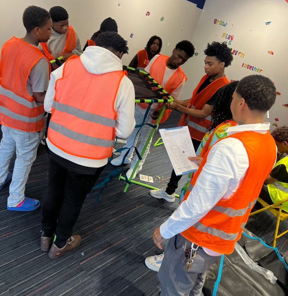 People wearing orange safety vests gather around a green metal frame while one person holds an instruction sheet and wall text reads “grow play learn together”