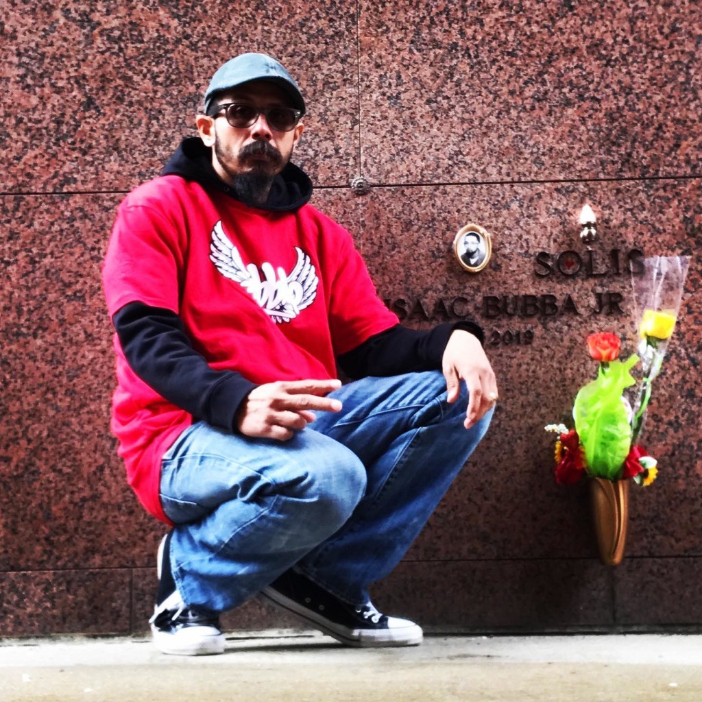 A man in a red shirt and cap crouches in front of a wall with an engraved name and a small arrangement of flowers nearby.