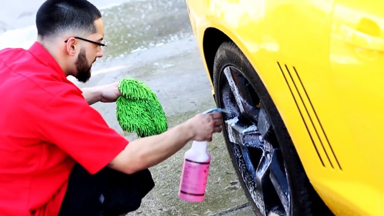 A man kneels next to a yellow car, a green umbrella in one hand and a spray bottle with soapy suds next to a wheel in the other.