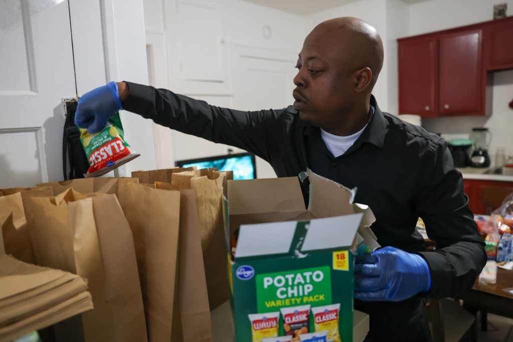A person wearing blue gloves places a bag of chips into a row of paper bags, with a box labeled 