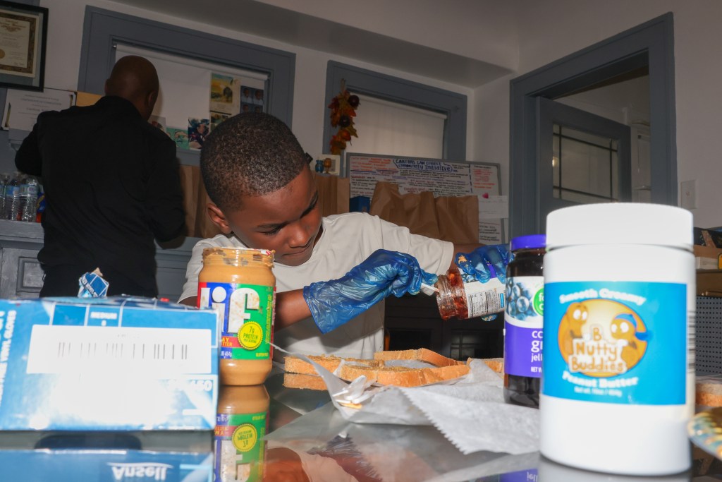 A person wearing blue gloves holds a utensil that is inside a jar of jelly at a table with jars labeled 