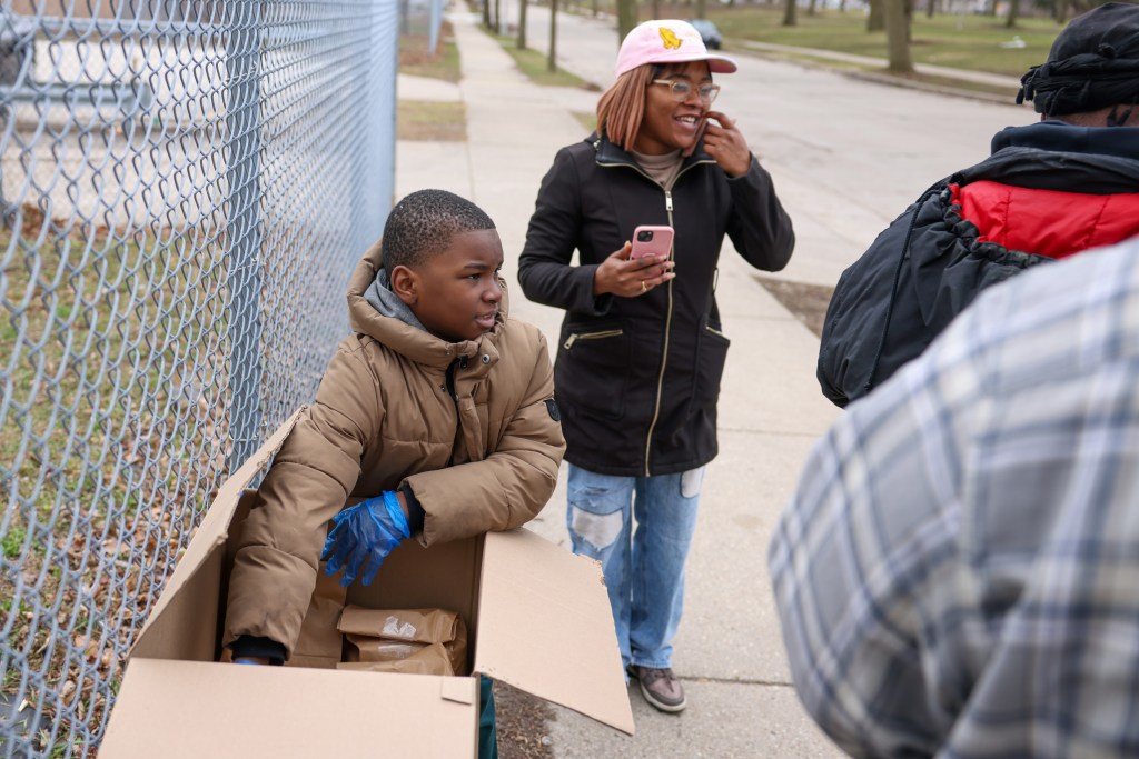 A person wearing a brown jacket and blue gloves reaches into a cardboard box on a sidewalk while another person holds a pink phone nearby, with a chain-link fence to the left, two other people partially seen on the right and a street in the background.