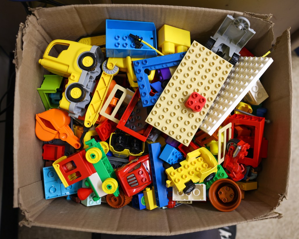 A cardboard box contains colorful building blocks, toy vehicle parts, wheels and base plates piled together.