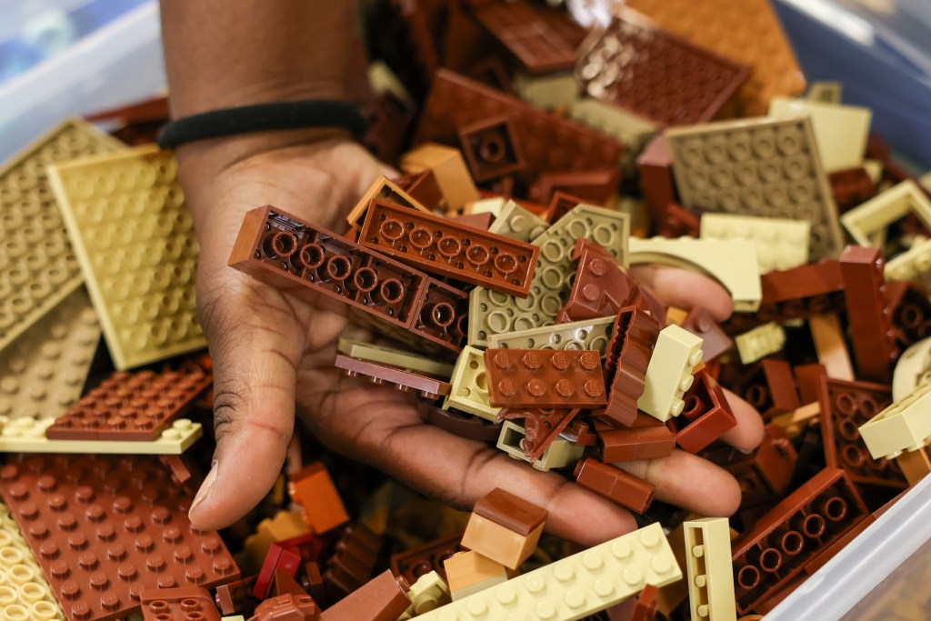A person's hand holds brown and tan building blocks above a bin filled with similar pieces.