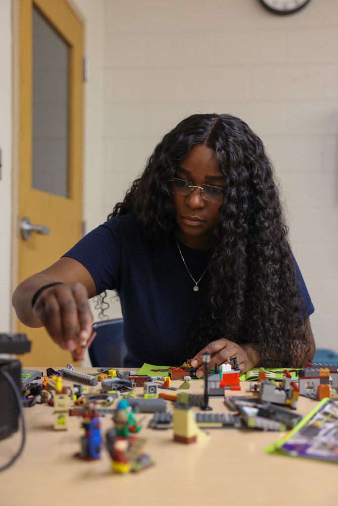 A person with long curly hair and glasses places a building piece on a table covered with assembled models and loose blocks in a room with a door and a clock partially visible.