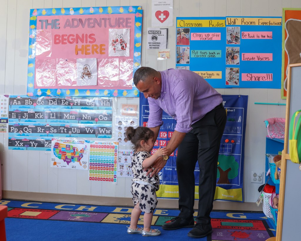A person in a purple shirt bends toward a child standing on a classroom rug. Colorful posters, alphabet charts and classroom rules are displayed on the wall behind them.