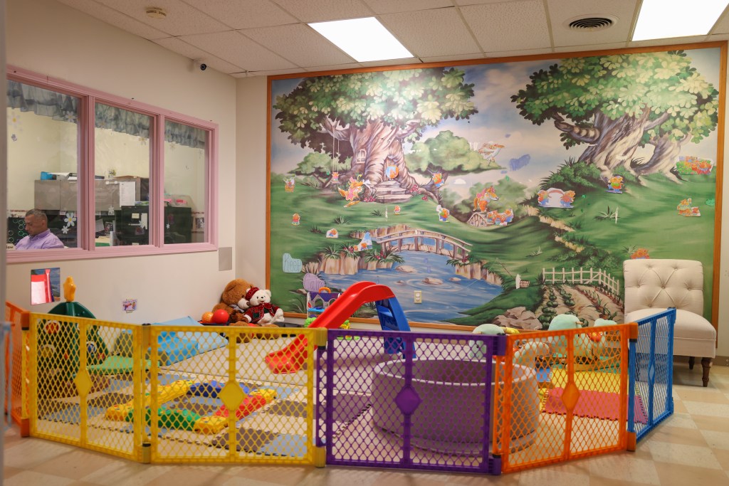 A fenced indoor play area contains toys, a small slide and mats. A wall mural shows trees, a stream and a bridge, and a person is visible through an interior window.