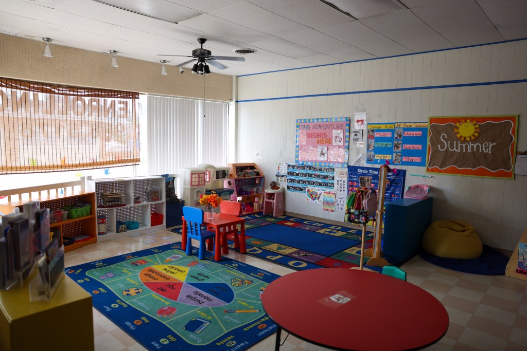 A classroom contains colorful rugs, child-sized tables, toys and educational posters. A bulletin board reads “Summer,” and another reads “THE ADVENTURE BEGNIS HERE.”
