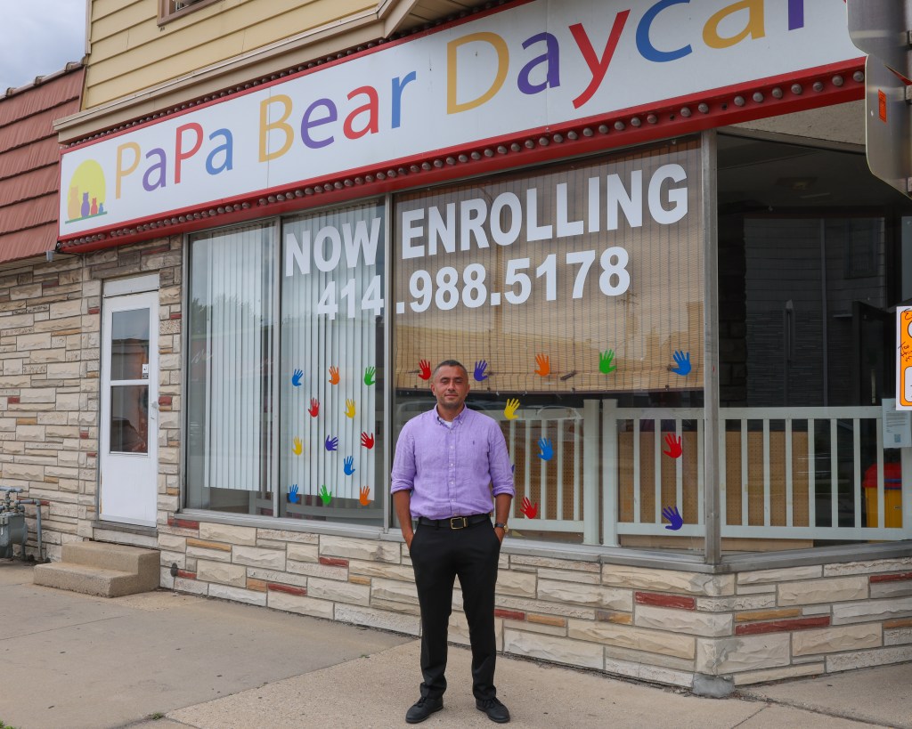 A person stands on a sidewalk in front of a building with a sign reading “PaPa Bear Daycare.” Large window text reads “NOW ENROLLING 414.988.5178.”