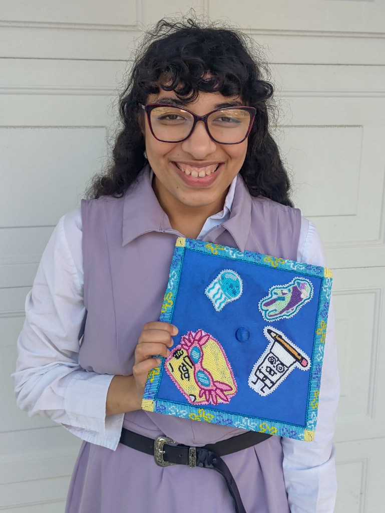 A person wearing glasses holds a blue beaded square featuring a fish, a medicine bottle, and other beadwork designs while standing in front of a garage door.