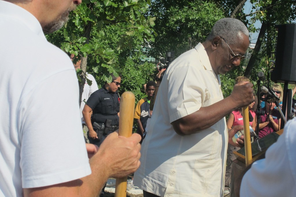 A person holds a long wooden handle outdoors surrounded by people. Trees, cameras and a uniformed officer are visible in the background.