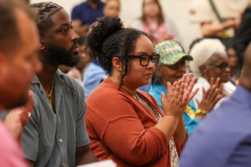 People sit in rows. One person wearing glasses applauds while others look to the right.