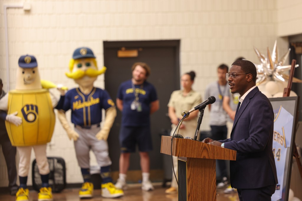A person speaks at a podium into a microphone. Baseball-themed mascots and a framed "Brewers" jersey with the number "44" stand in the background.
