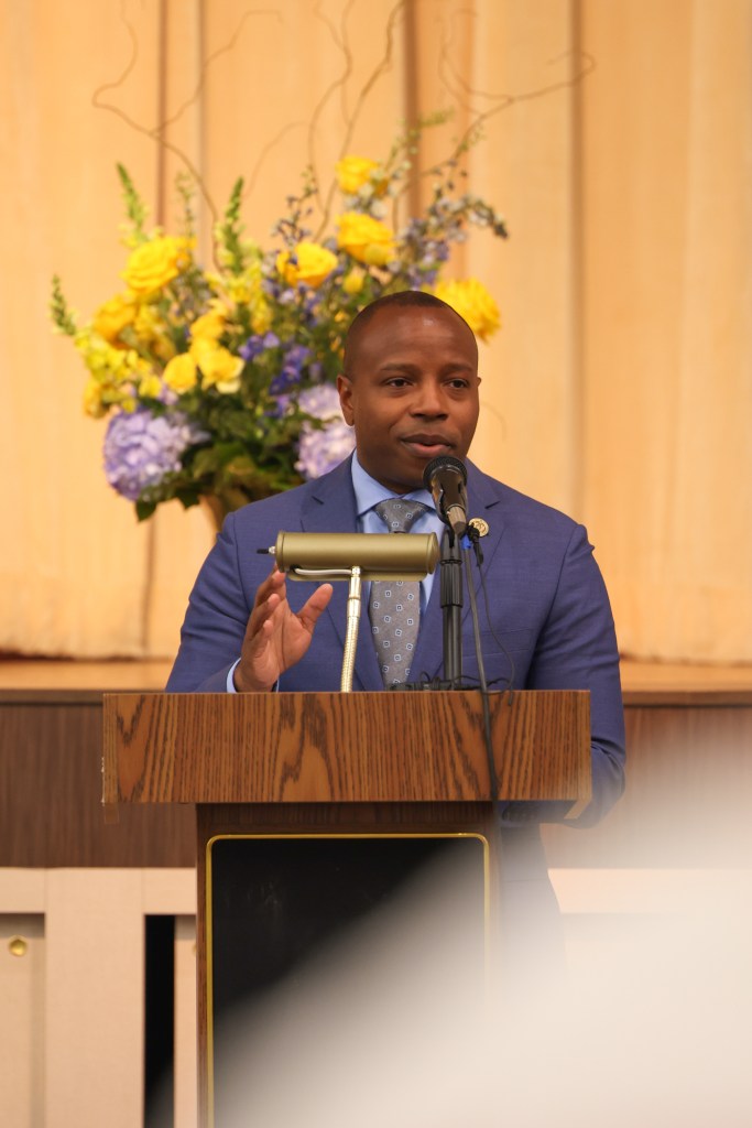 A person in a blue suit speaks at a podium into a microphone. A large floral arrangement stands behind the podium.